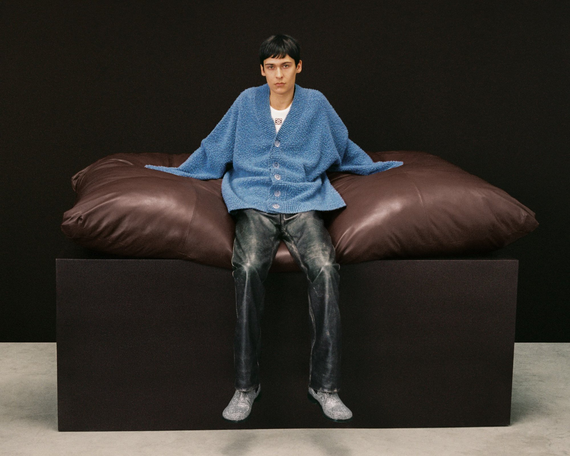 A slender adult poses indoors wearing a blue cardigan, grey jeans, and silver shoes, seated on a large brown pillow on a black block.