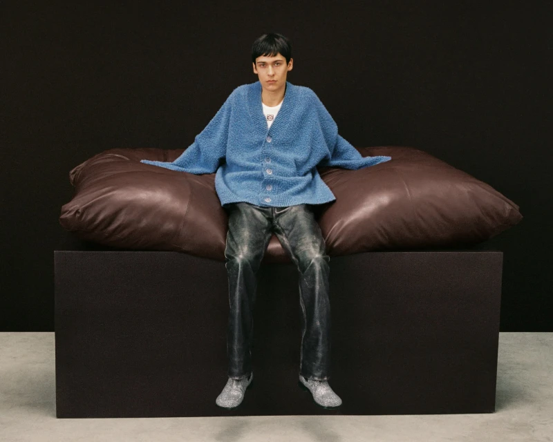 A slender adult poses indoors wearing a blue cardigan, grey jeans, and silver shoes, seated on a large brown pillow on a black block.