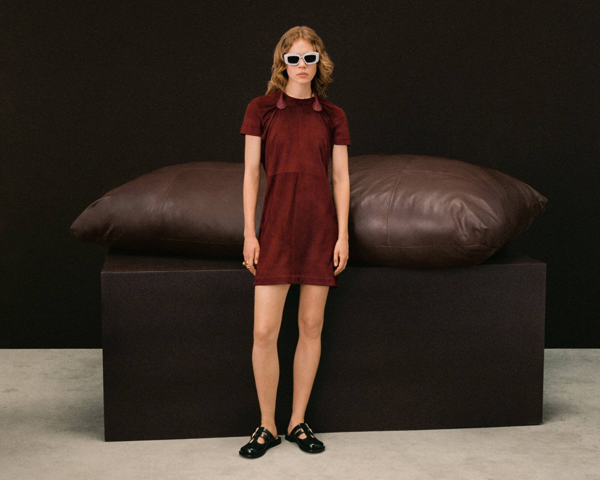 Full-shot of a fashionable woman wearing a burgundy dress and sunglasses, standing in front of a black backdrop and brown leather pillow.