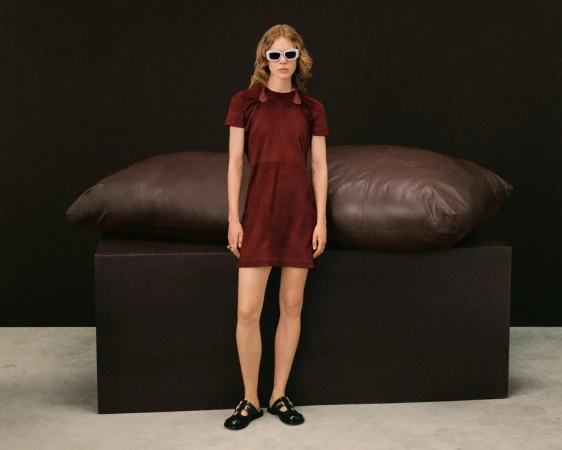 Model wearing a dark red dress and white sunglasses standing in front of oversized leather cushions on a dark block.