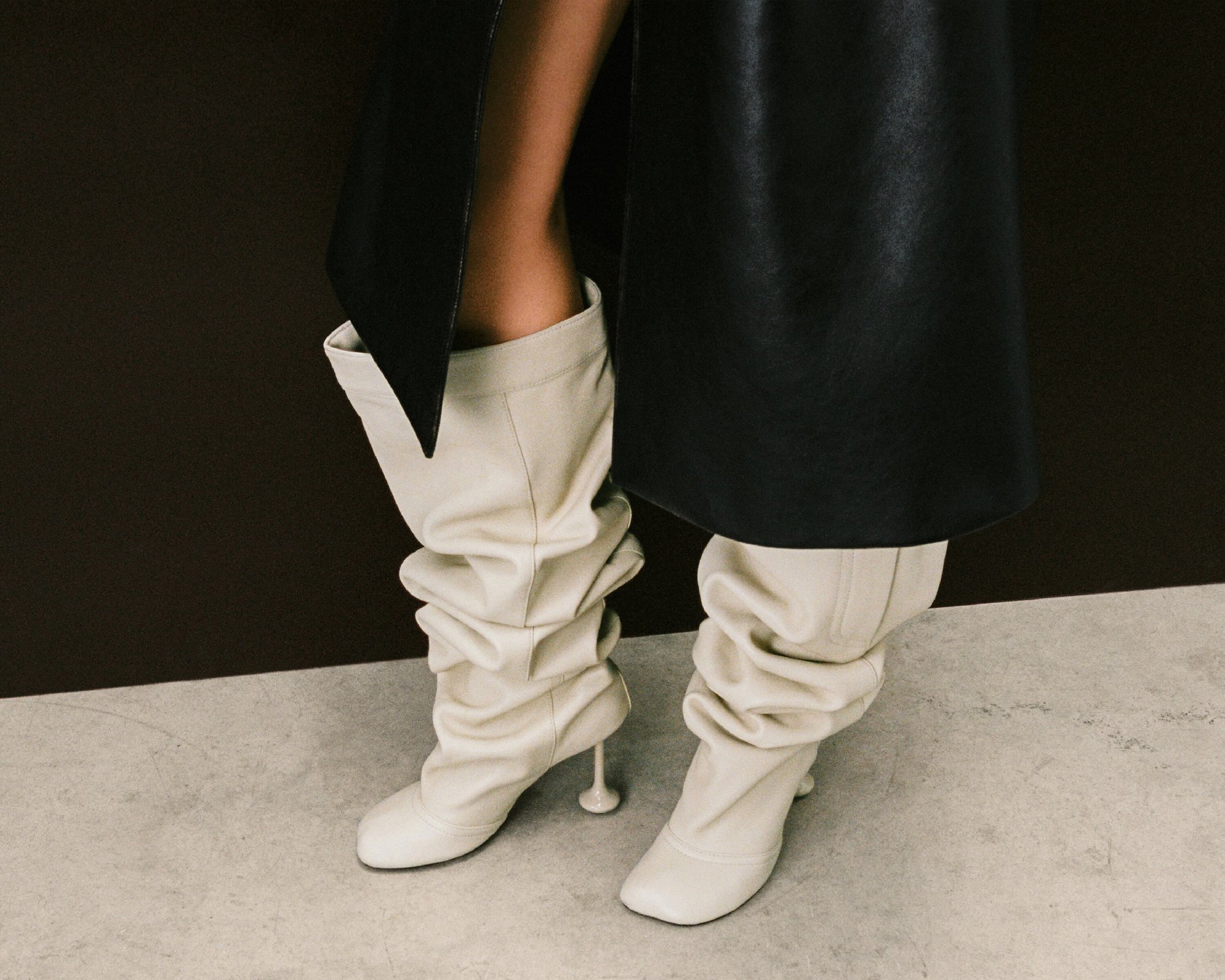 Pair of slouchy, ivory-colored leather boots with a distinct heel design. The boots are set against a dark brown and grey background.