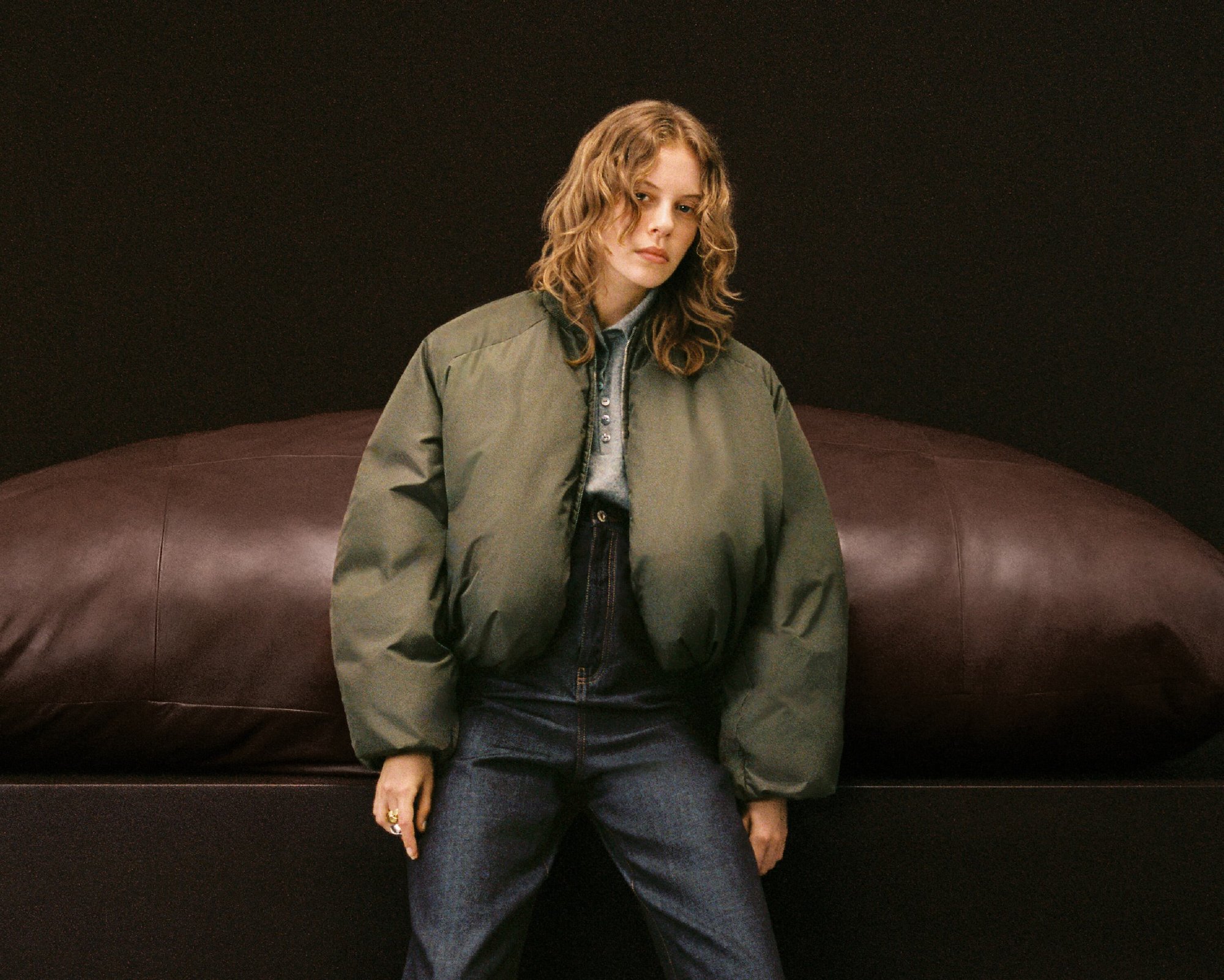 A young woman with wavy hair poses indoors in an olive puffer jacket, blue jeans, and a button-down shirt against a dark backdrop.