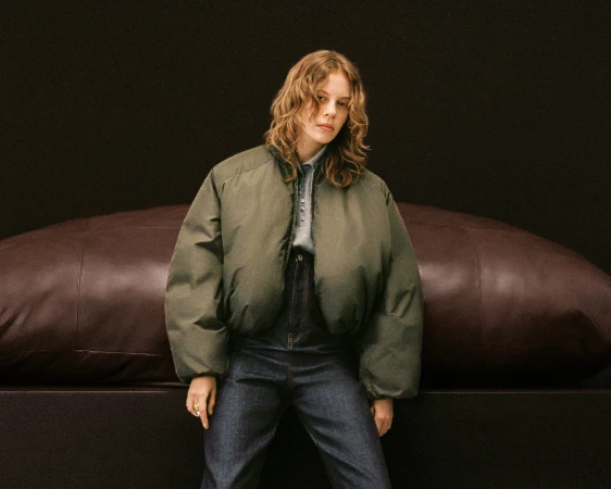 A young woman with wavy hair poses indoors in an olive puffer jacket, blue jeans, and a button-down shirt against a dark backdrop.