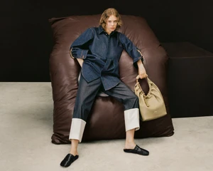 Model seated on a large brown leather beanbag wearing a denim shirt and trousers, holding a beige shoulder bag.