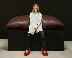 Model seated on a dark block wearing a white tunic, black trousers, and textured red shoes against a dark background