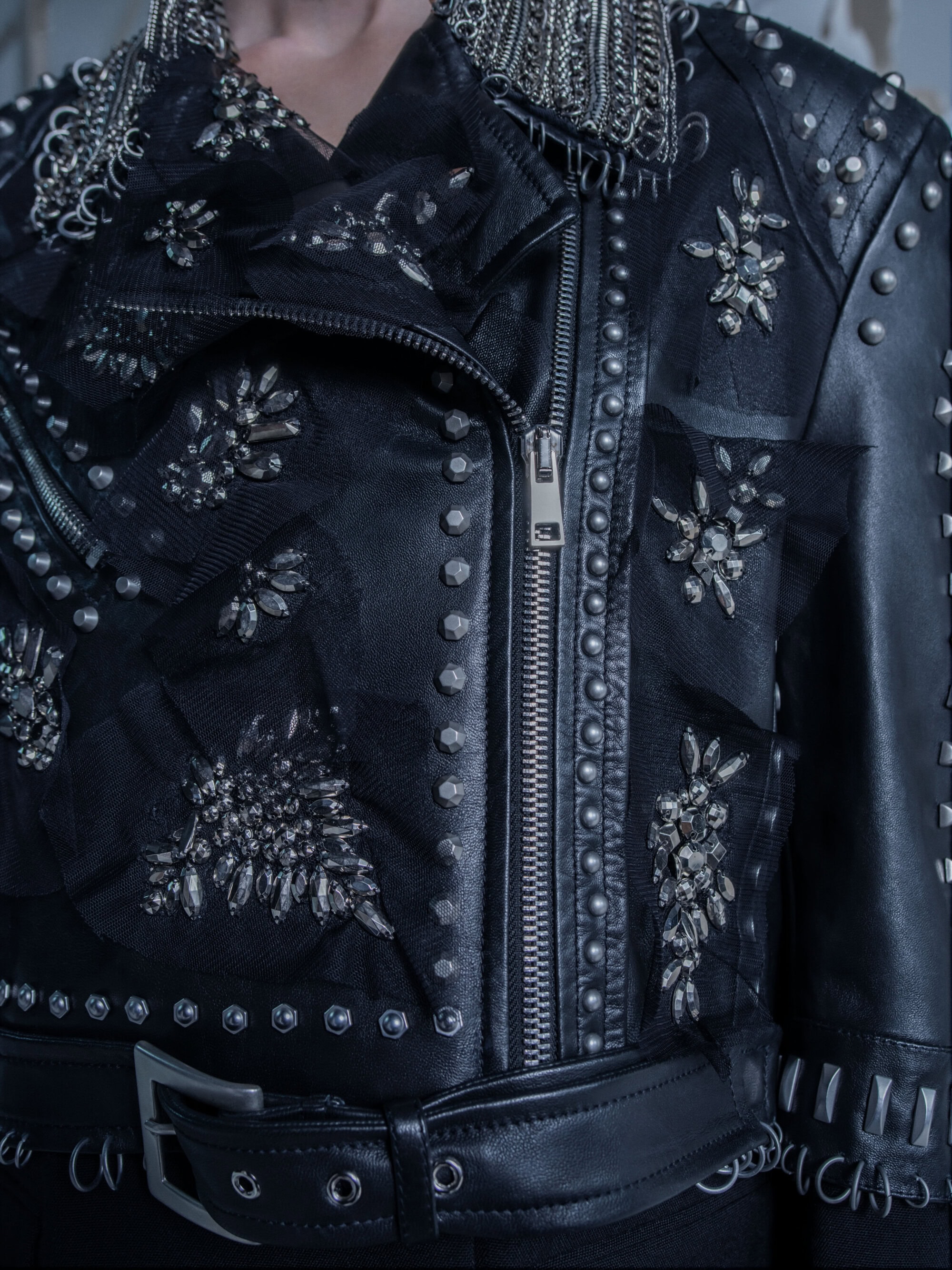 Close-up of a black leather jacket with silver studs, rhinestones, zipper, and a belt, accessorized with metal chains and sheer fabric.
