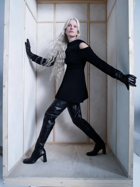 A slender woman with long white hair and black leather boots poses in a wooden box wearing a black dress against a bright studio background.