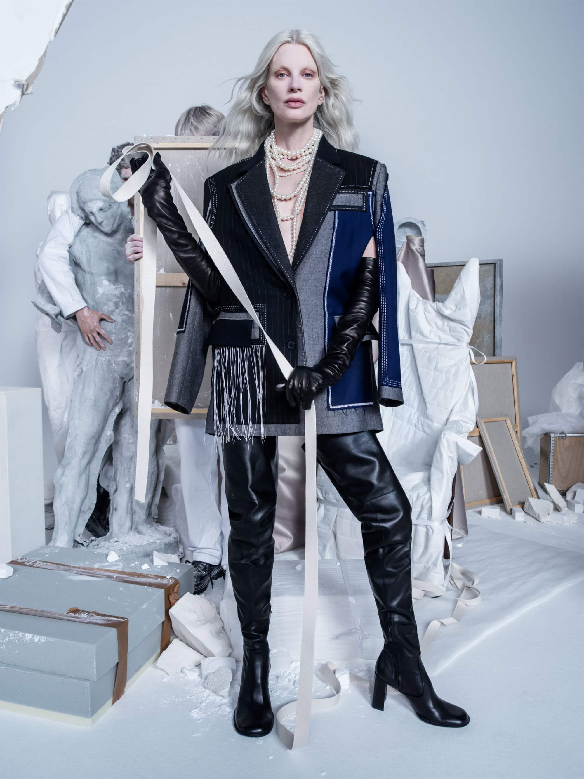Fashion editorial shot of a slender model in a deconstructed blazer, pearl necklaces, leather gloves and thigh-high heeled boots.