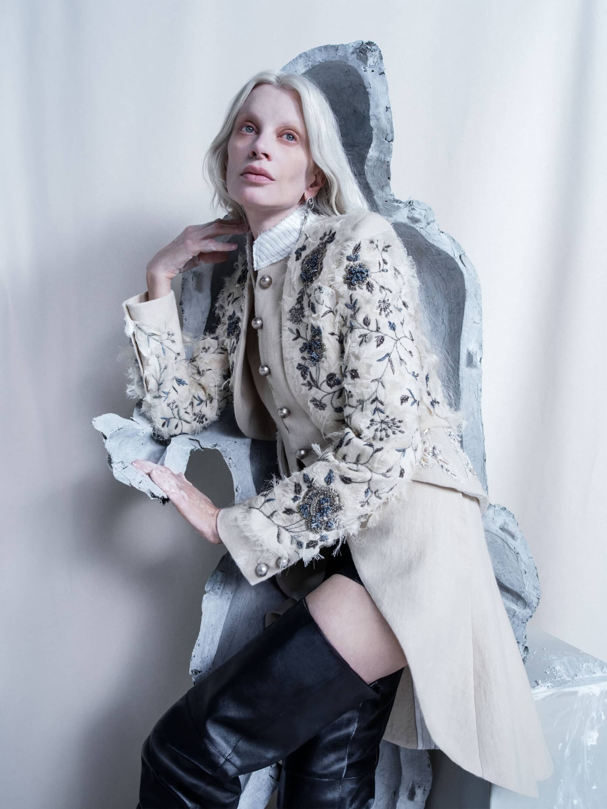 Woman with platinum hair in an embroidered jacket poses next to concrete blocks in a studio portrait with soft lighting.