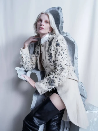 Woman with platinum hair in an embroidered jacket poses next to concrete blocks in a studio portrait with soft lighting.