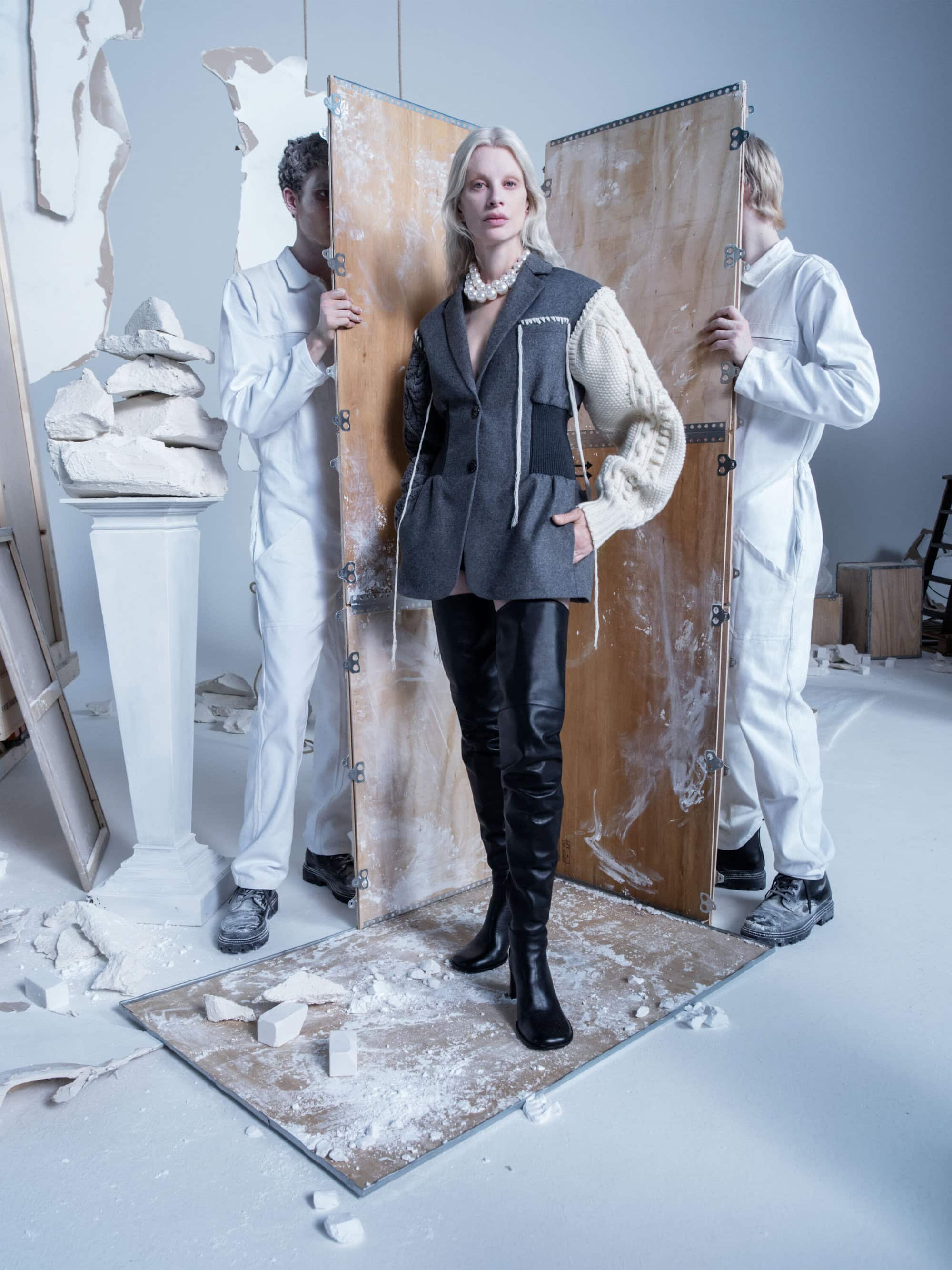 Fashion editorial shot of a model in an asymmetrical blazer and thigh-high boots posing between two men in white coveralls in a studio setting.