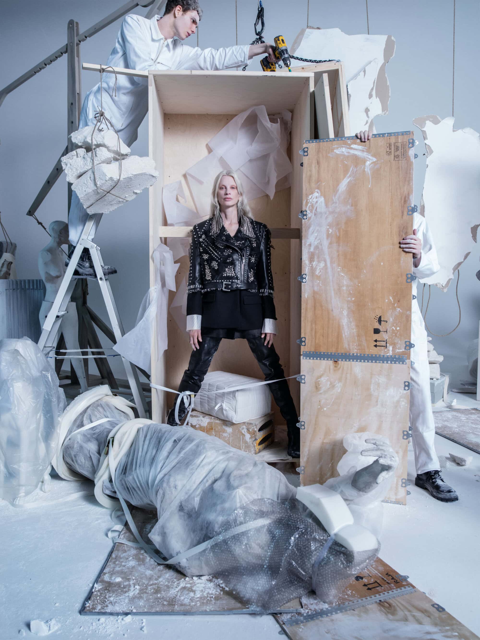 Fashion-forward editorial shot of a woman in a studded jacket standing amid sculptures in an art studio setting with two men in work coats.