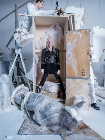 Fashion-forward editorial shot of a woman in a studded jacket standing amid sculptures in an art studio setting with two men in work coats.