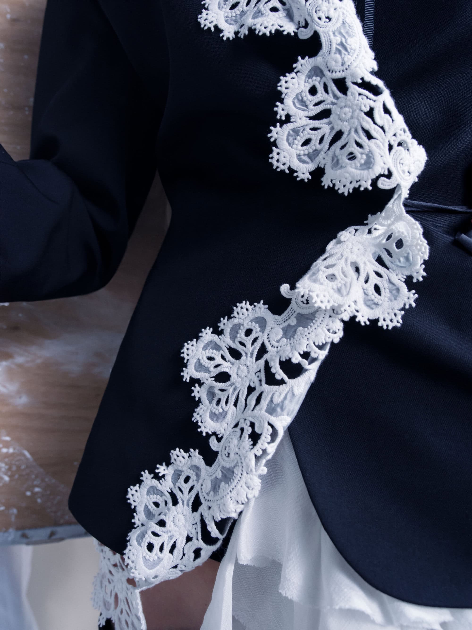 Close-up of a dark blue jacket trimmed with intricate white lace worn over a white ruffled skirt.