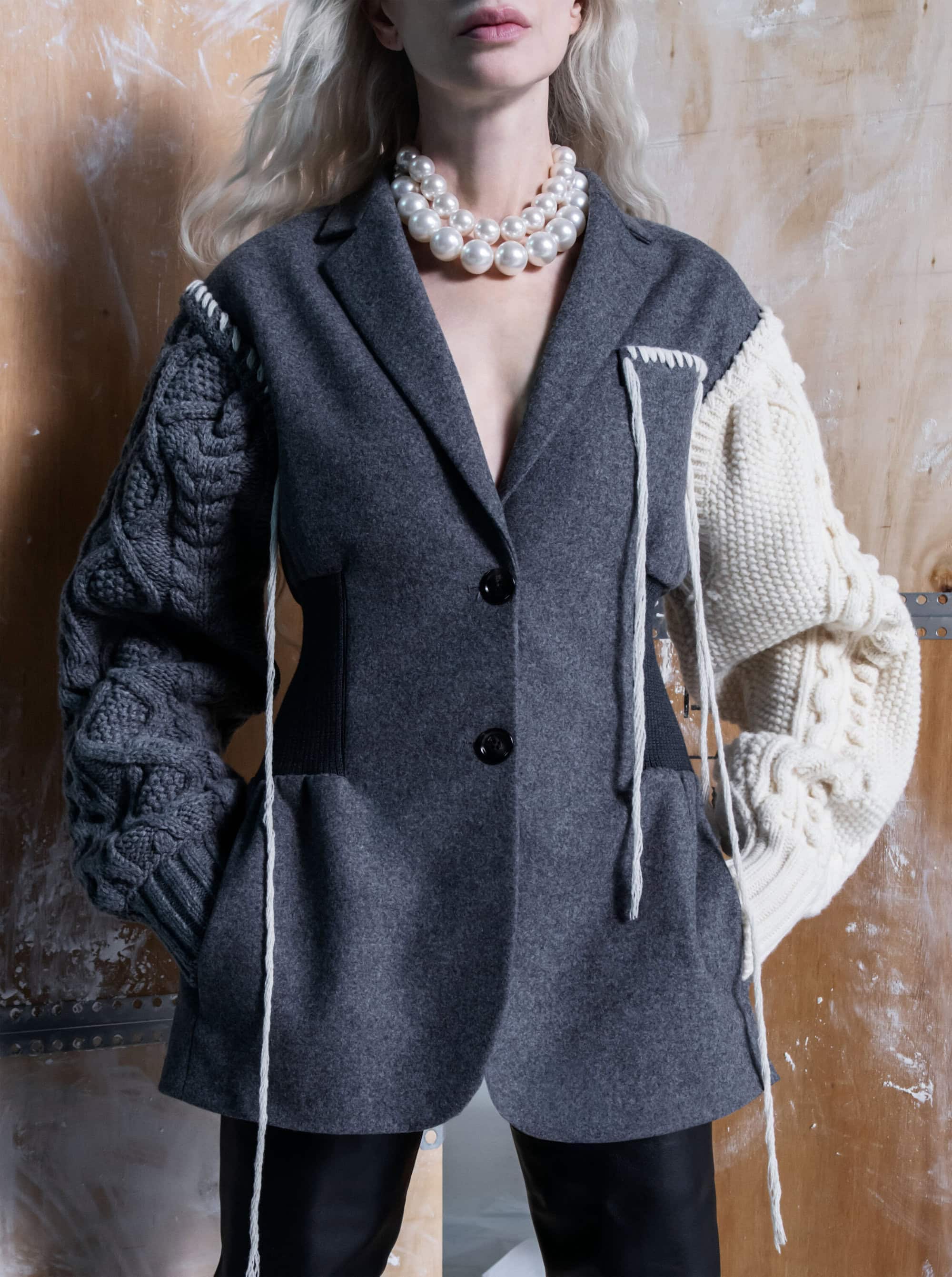 A fair-skinned person models a gray blazer with cable-knit sleeves, wearing a pearl necklace against a textured wood background.