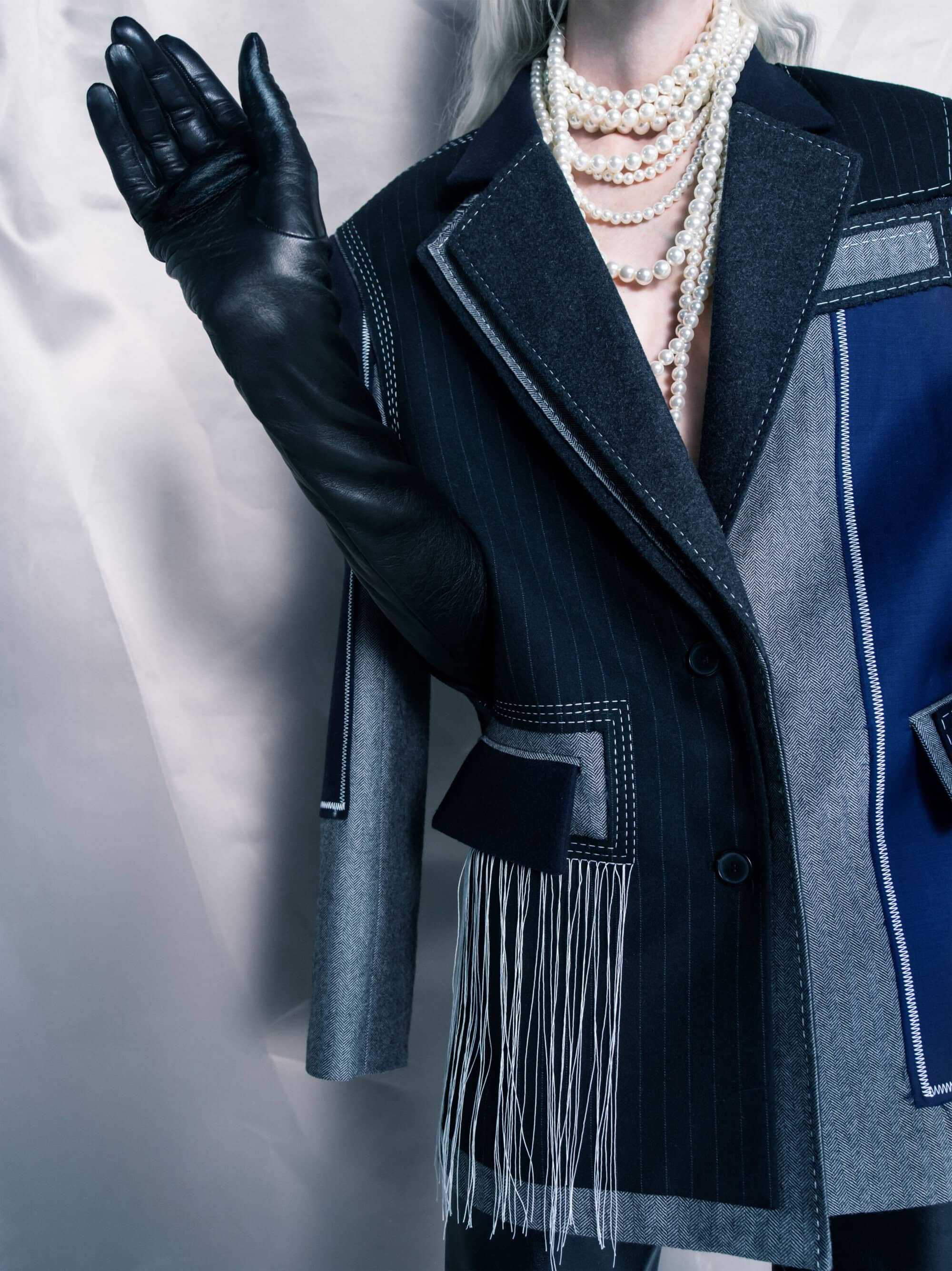 Woman wearing a pinstripe patchwork blazer with leather gloves and pearl necklaces against a soft grey satin background.