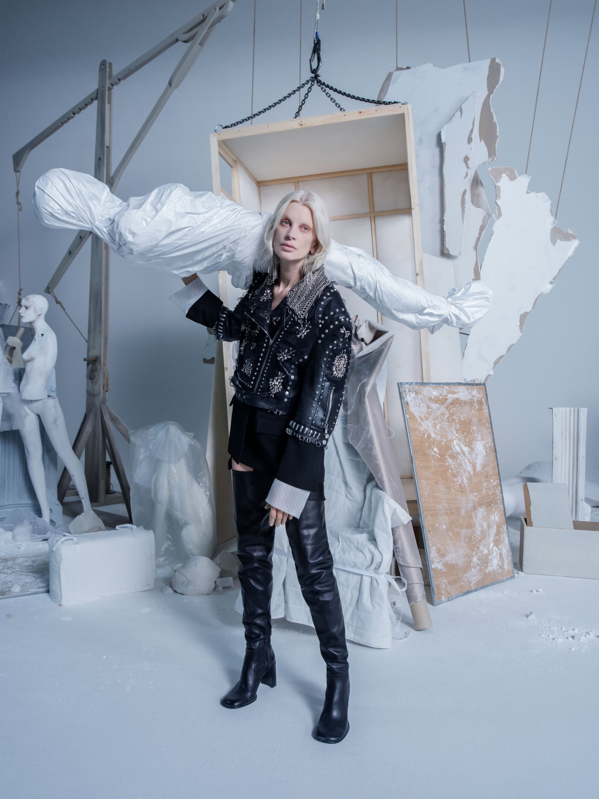 Fashion-forward blonde woman in black leather jacket posing with a mannequin piece in a light studio with art props and sculptures.