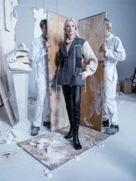 Model Kristen McMenamy wearing a grey blazer and over-the-knee black boots posed between wooden partitions in a studio by Tim Walker.