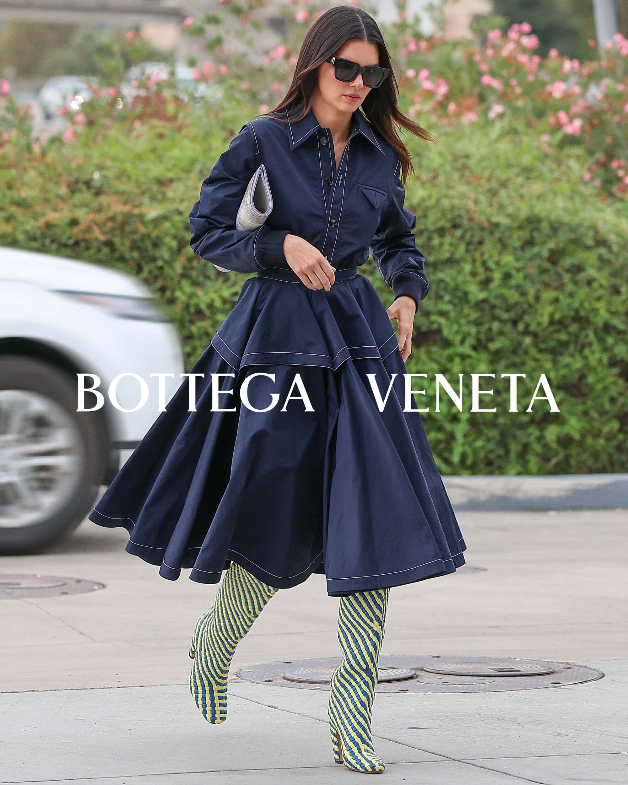 A woman in a blue dress and patterned boots walks on a street, carrying a light grey purse, with a white car and greenery nearby.