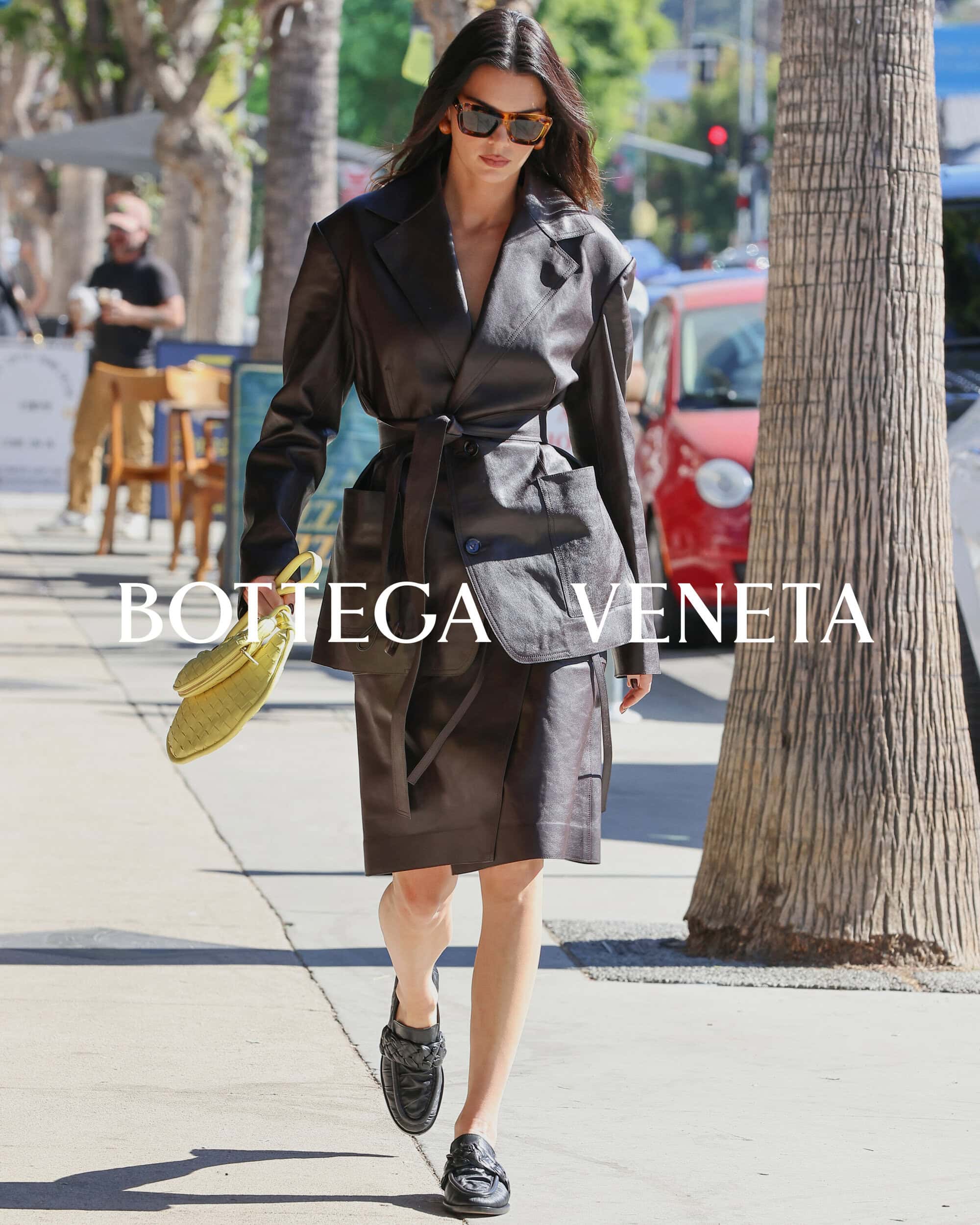 Woman in dark brown blazer dress with a matching purse and black loafers, walking on a city street on a sunny day.