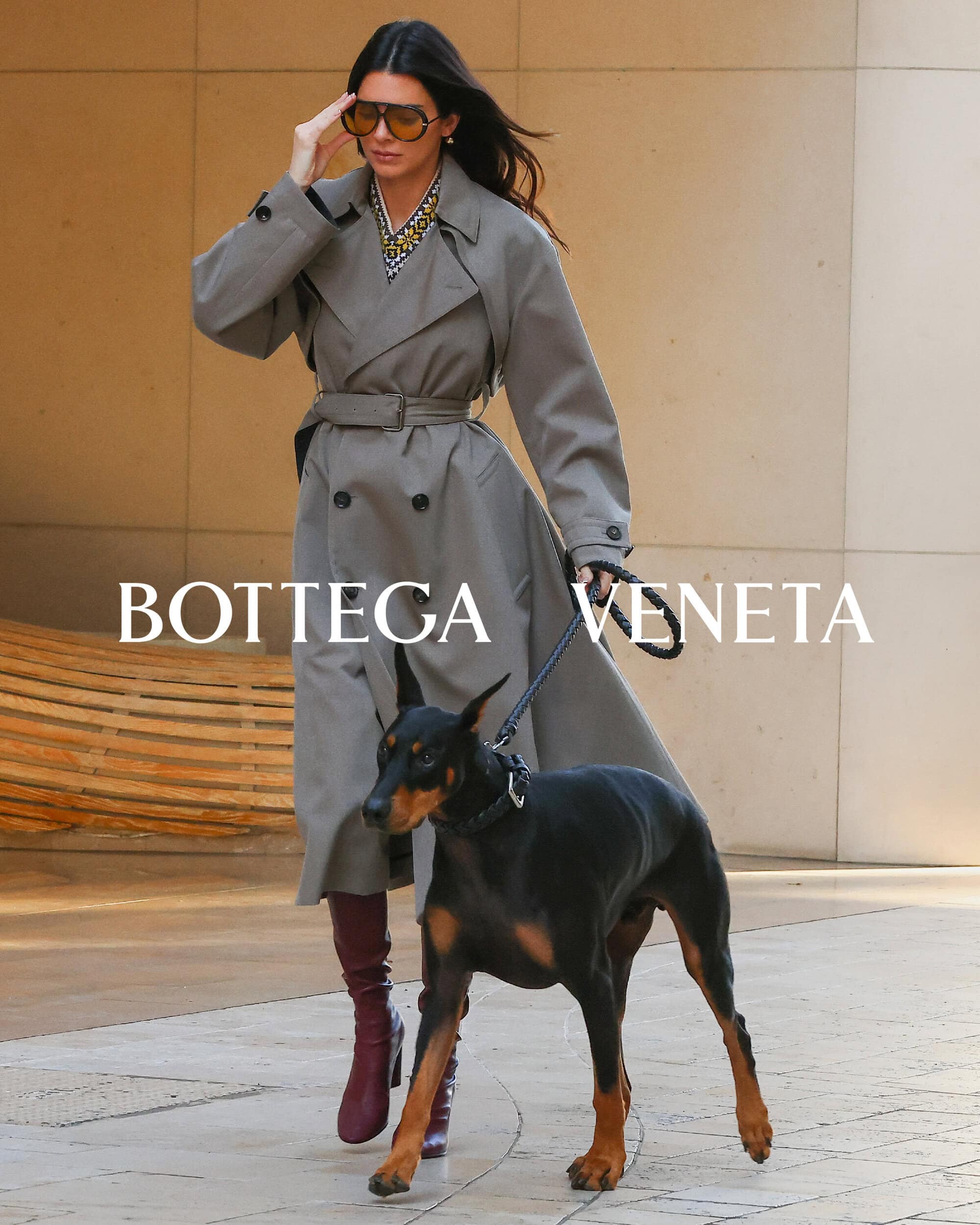 Adult female in an olive trench coat and sunglasses walks a black Doberman Pinscher dog on a leash. Bottega Veneta logo. Fashion shot.