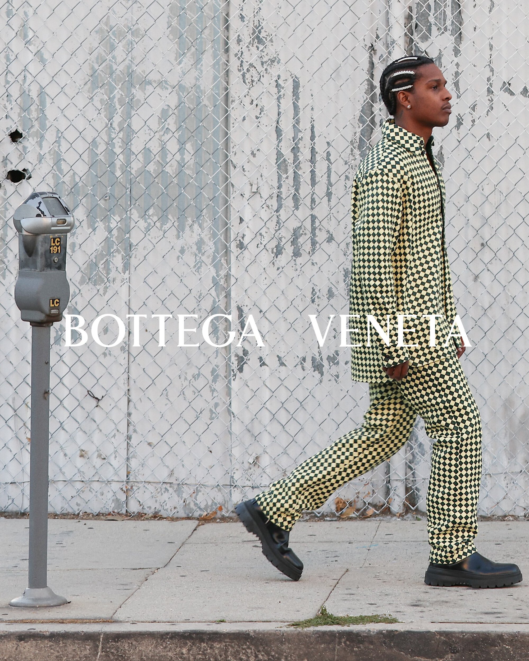 A male model walks along a city sidewalk wearing a patterned Bottega Veneta suit with a gray fence in the background.