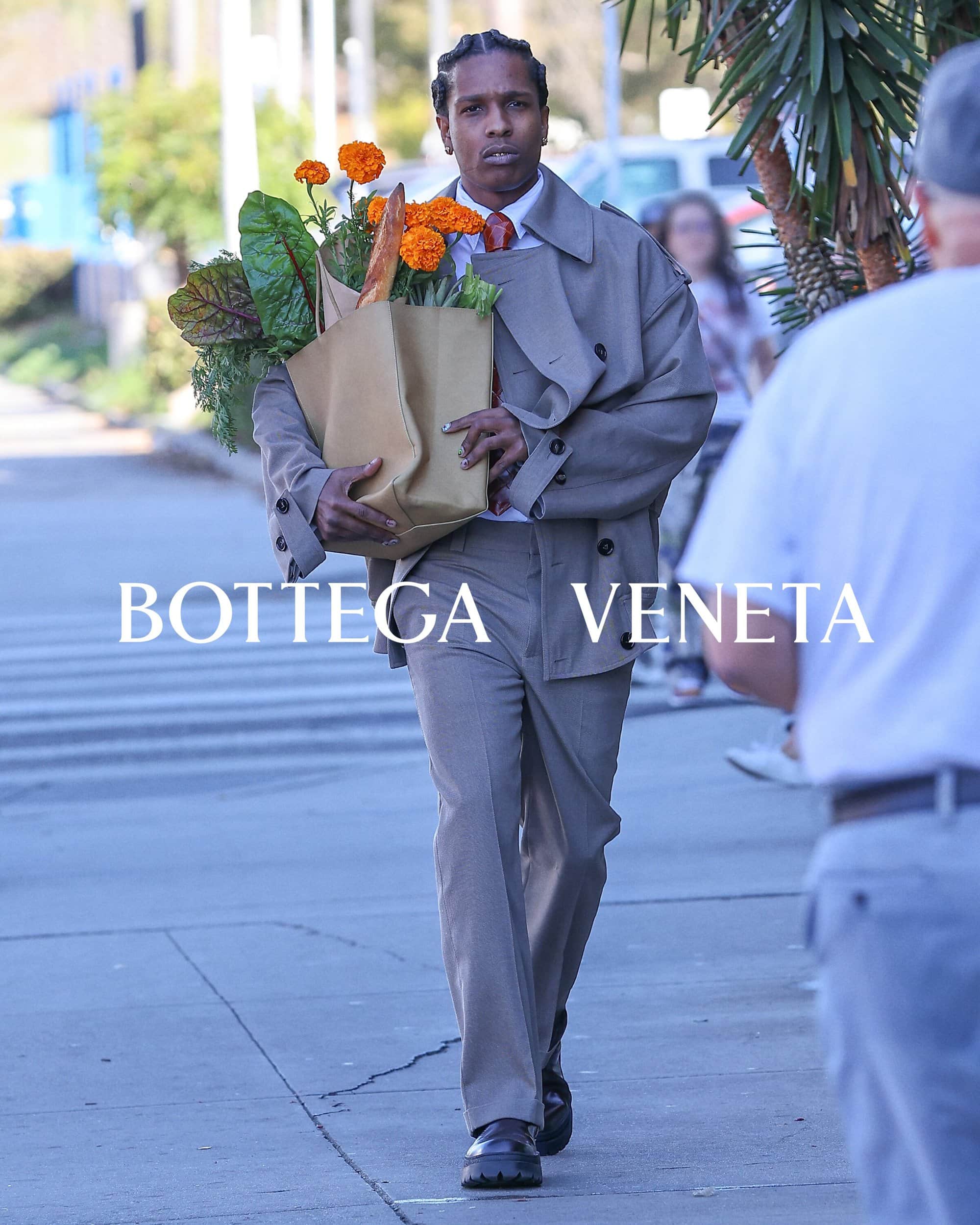 A$AP Rocky carries a bag of flowers and produce while walking on the sidewalk in a tan suit, captured in a medium shot.