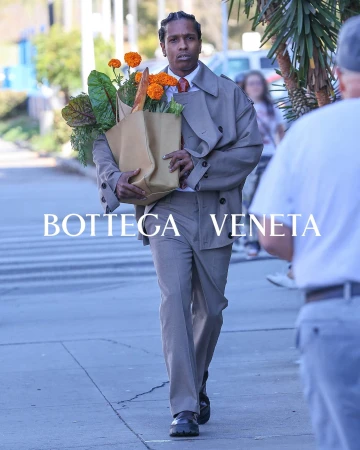 A$AP Rocky carries a bag of flowers and produce while walking on the sidewalk in a tan suit, captured in a medium shot.
