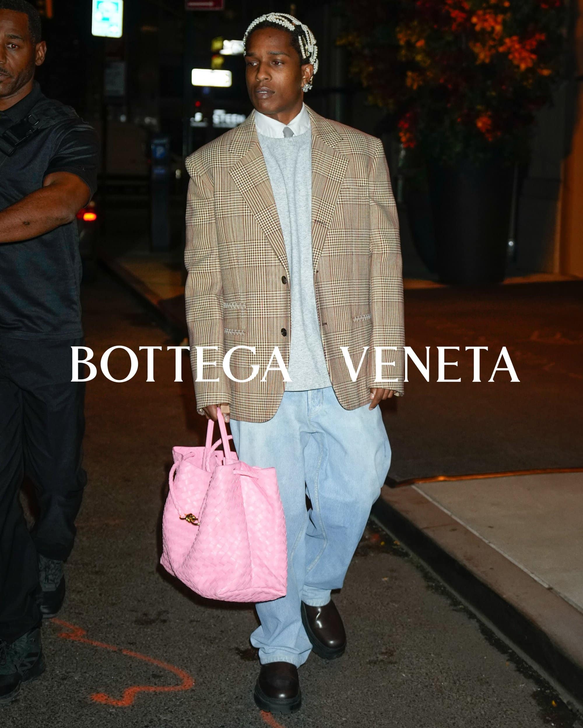 A$AP Rocky, an adult male, is walking on a city street with a pink Bottega Veneta purse and a plaid jacket over a gray sweater.