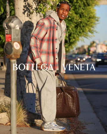 An adult male stands on a city sidewalk next to a parking meter wearing a grey sweatsuit, flannel shirt, and sneakers carrying a brown leather bag.