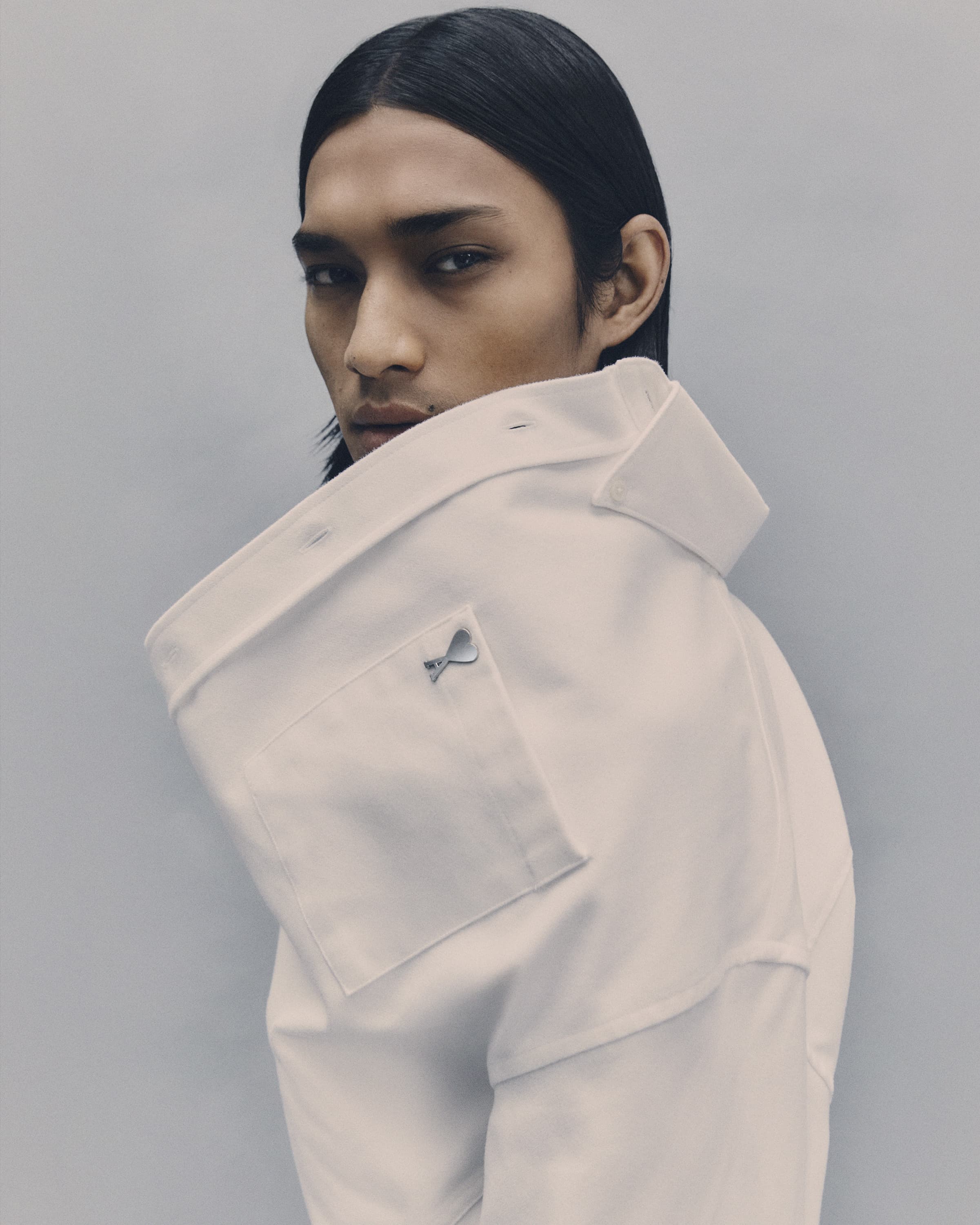 Portrait of an adult male with long dark hair, gazing at the camera, wearing a white collared shirt in a neutral studio setting.