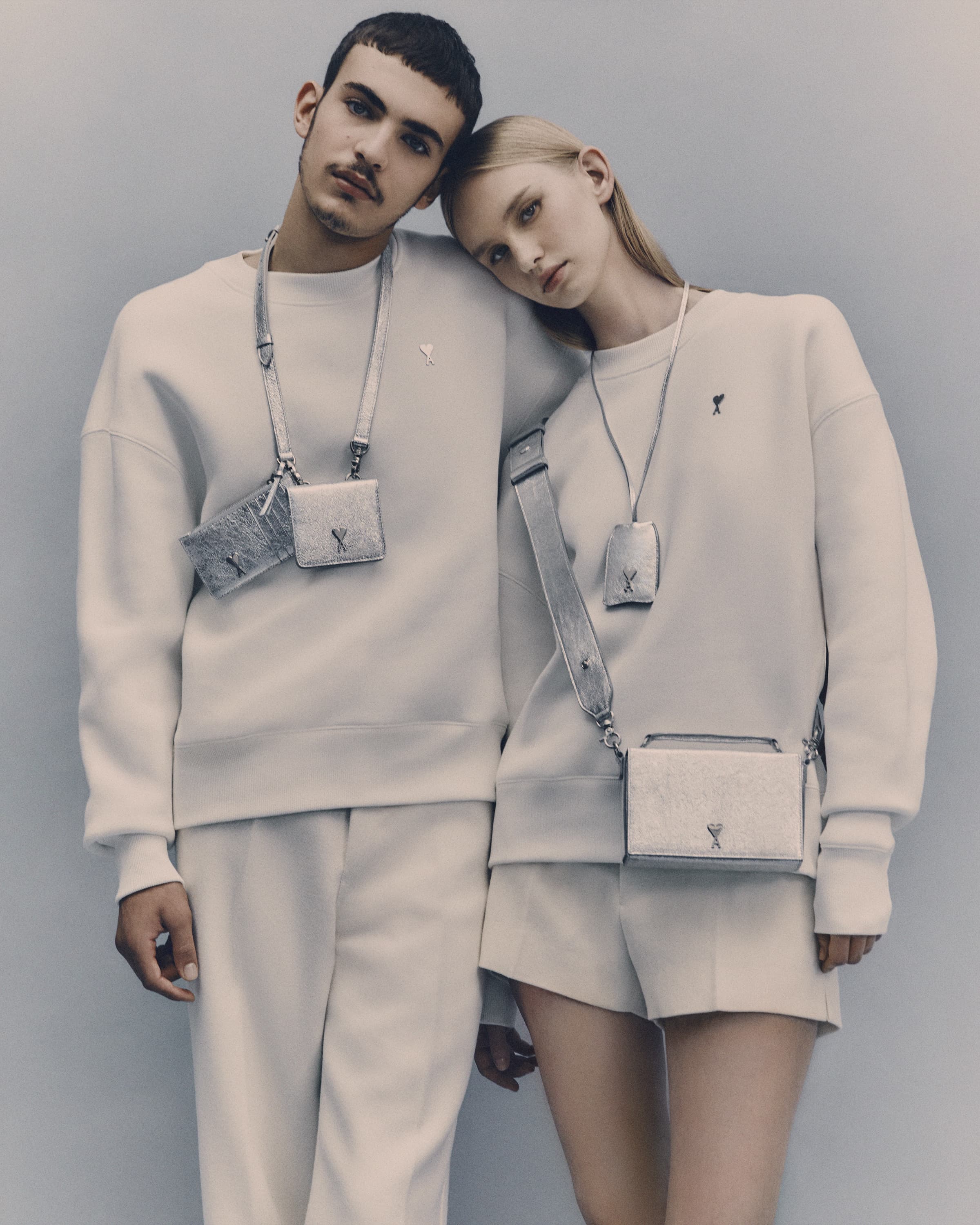 Fashion shot of a young adult male and female couple in matching off-white outfits with crossbody bags against a plain studio backdrop.