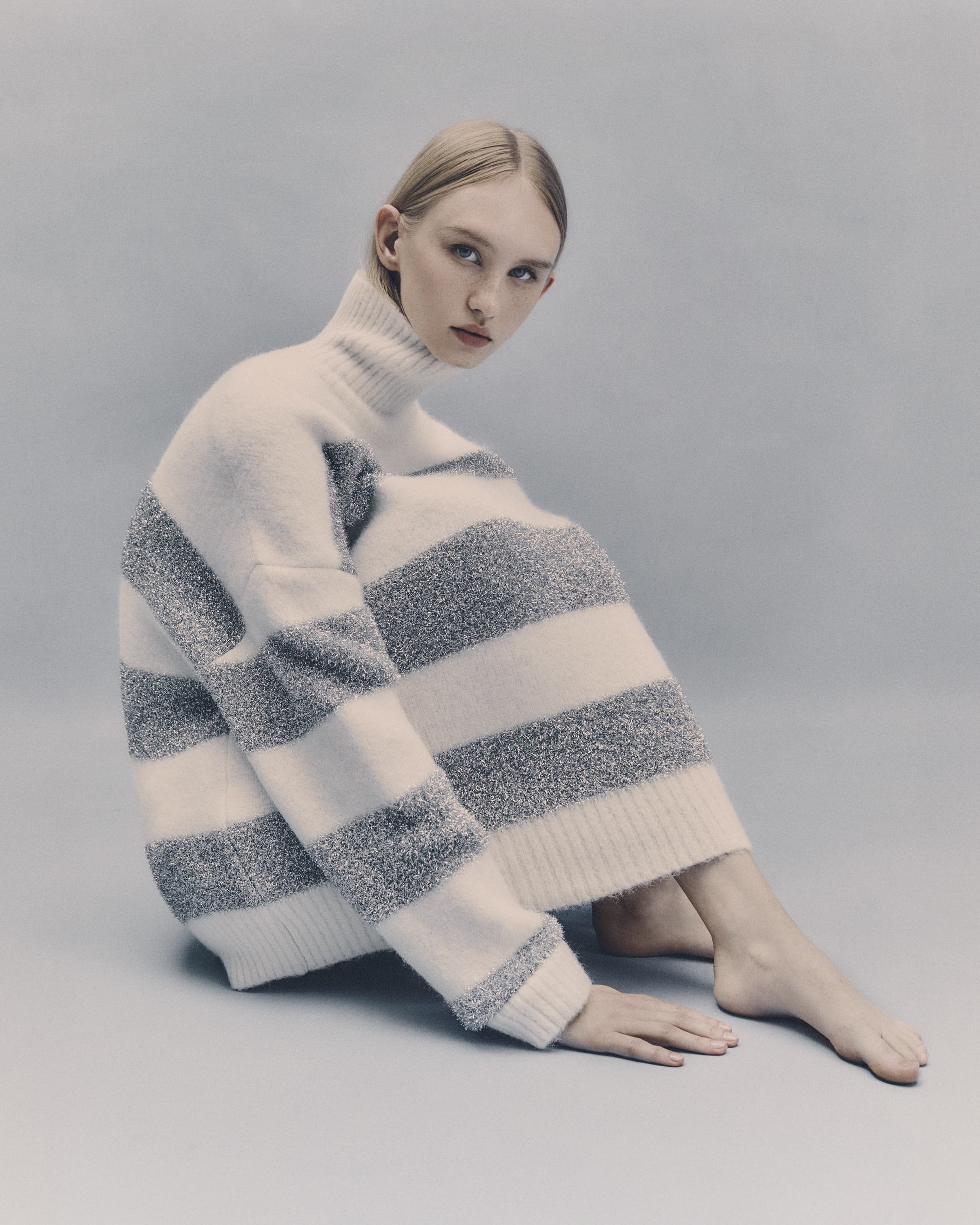 A fair-skinned young woman with blonde hair sits on a gray surface, wearing a striped sweater dress, gazing towards the camera in a studio setting.