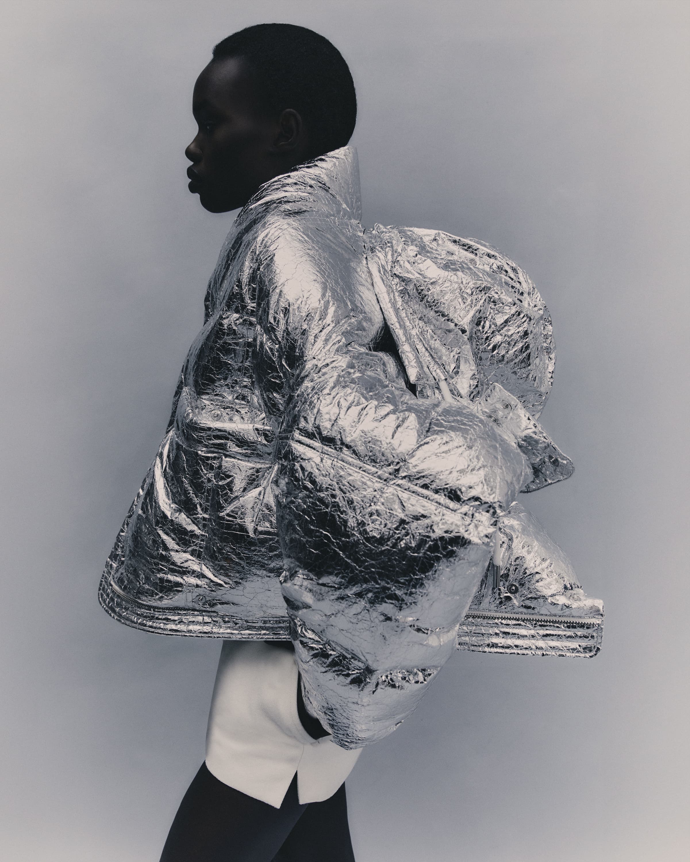 A dark-skinned fashion model poses in profile wearing a large metallic silver puffer jacket, short white skirt, and black stockings against grey.