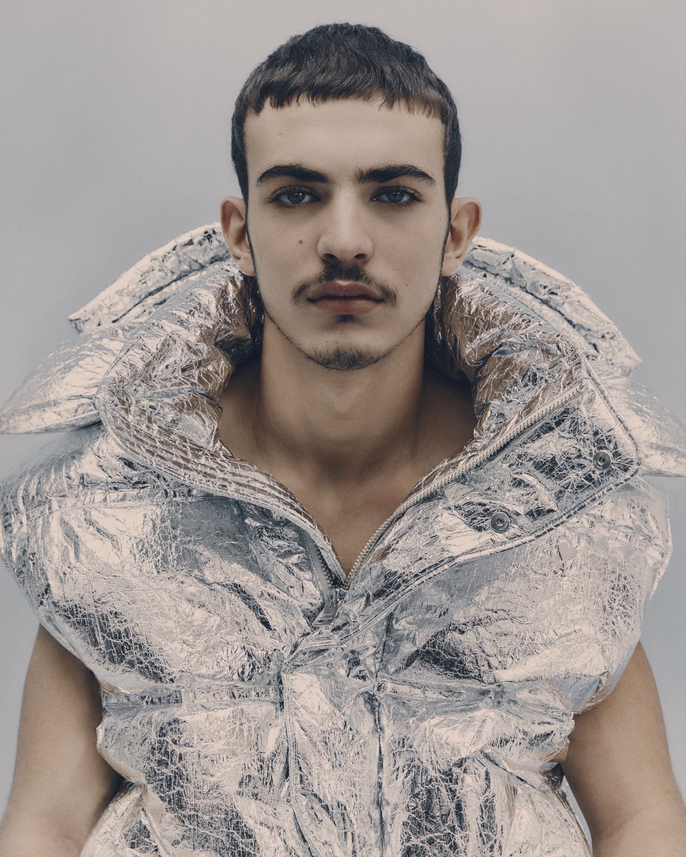 Close-up studio shot of a young adult male with a moustache wearing a shiny metallic silver puffer vest against a solid grey backdrop.