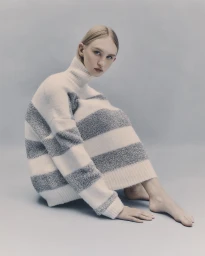 Model with blonde hair seated wearing a striped turtleneck sweater against a grey studio backdrop.