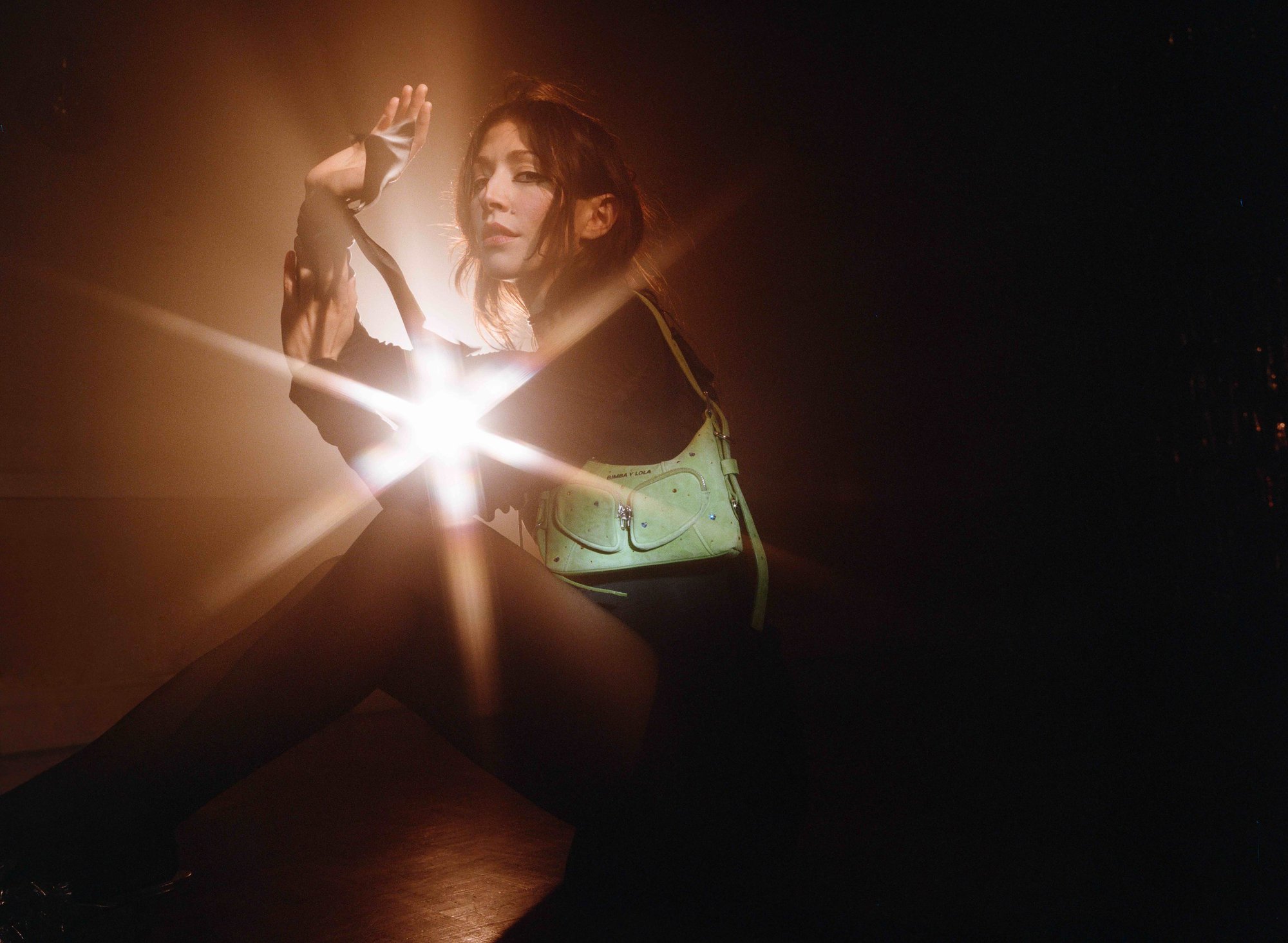 Portrait of a brunette woman sitting indoors in darkness, wearing a long-sleeve shirt, with a light green purse over her shoulder.