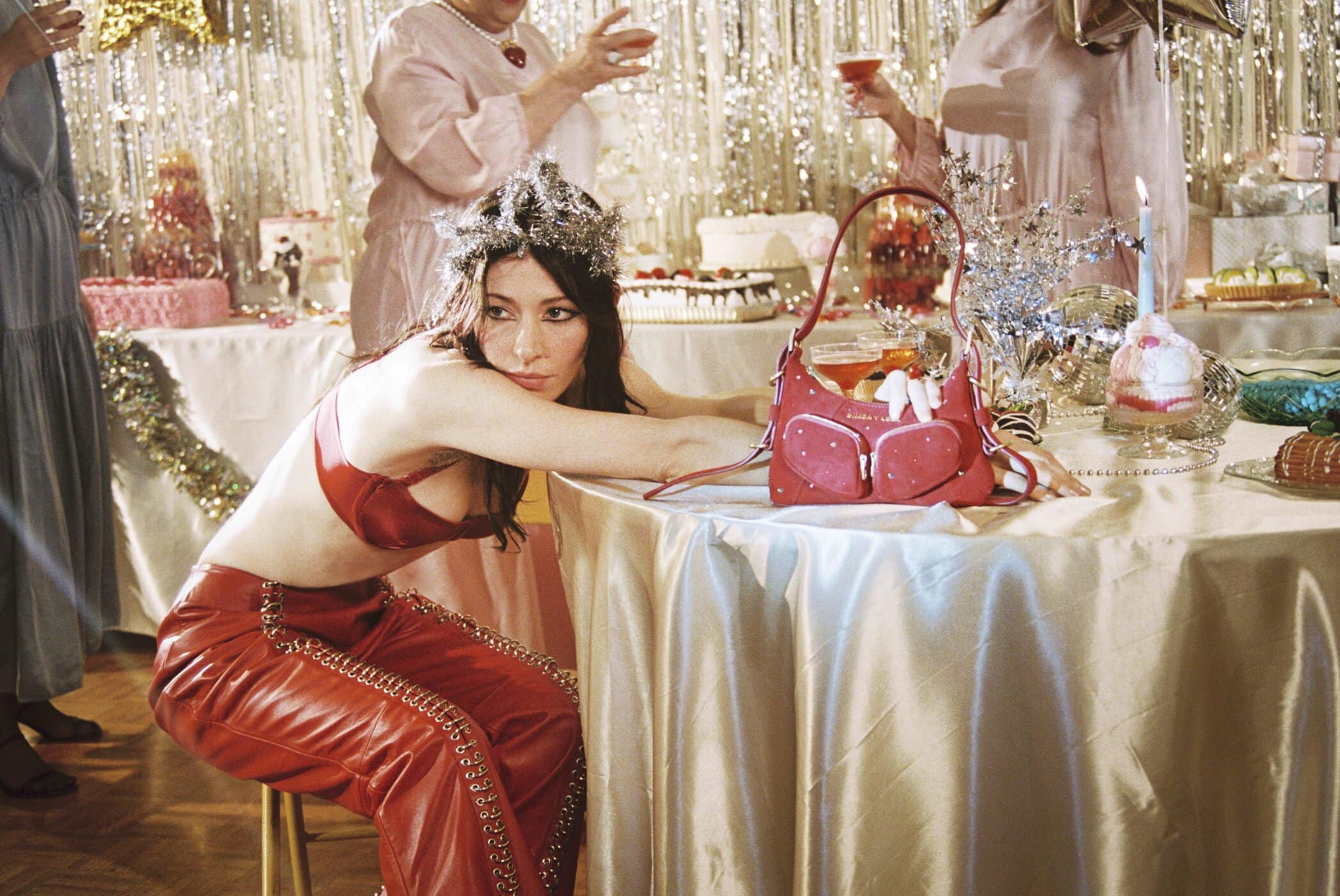 A stylish brunette woman in a red bra and pants sits at a banquet table laden with cake, drinks, and party favors. Tinsel backdrop.
