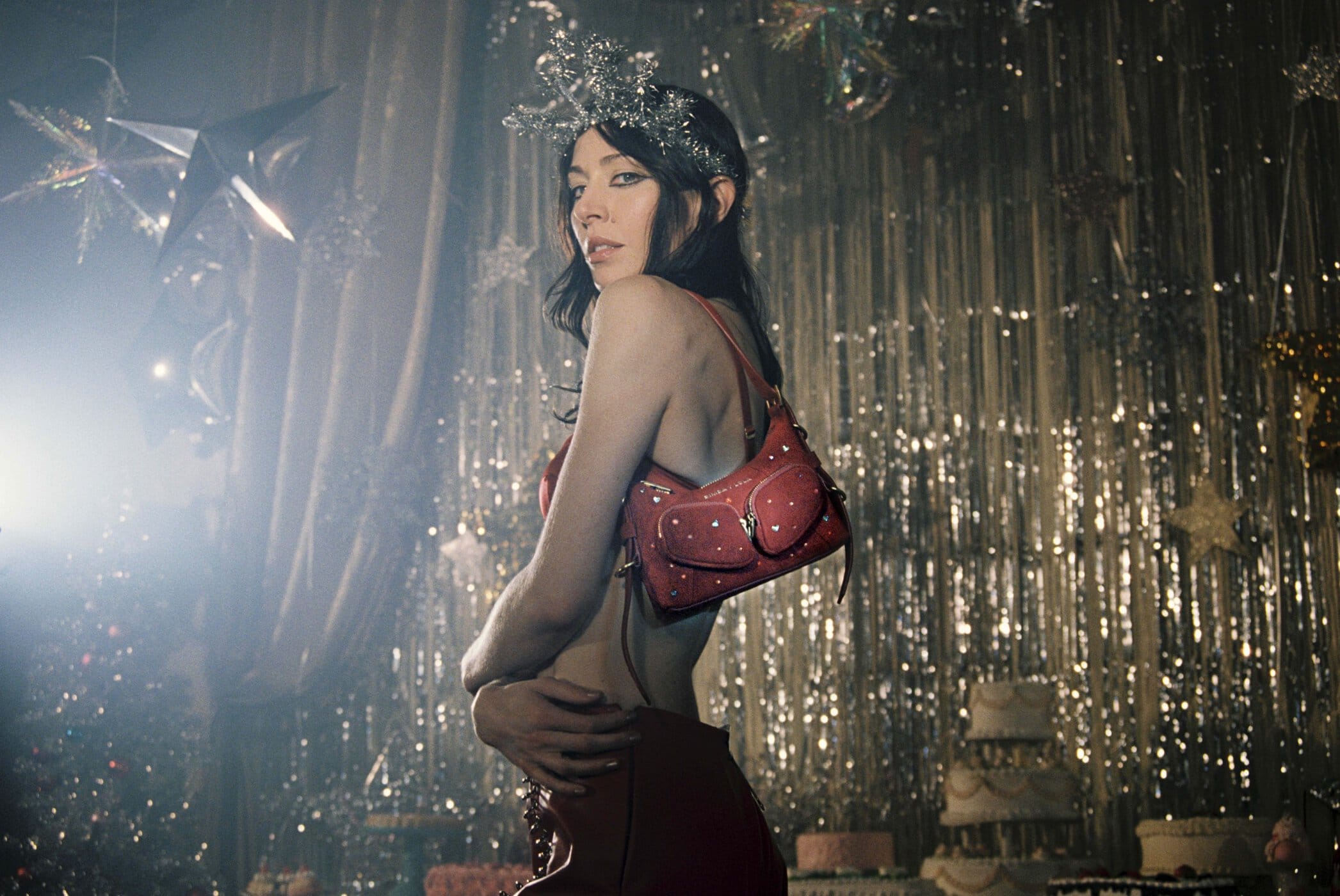 Woman with dark hair wearing a silver crown and red bag with stars. Background features glittery tinsel and tiered cakes.