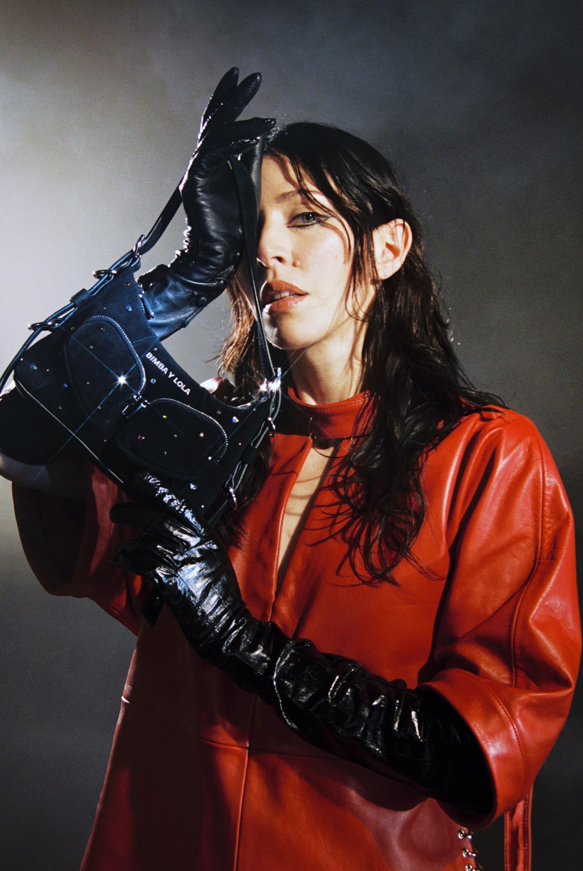A fashionable woman with long dark hair wearing a red dress and black leather gloves poses with a shiny black handbag in a studio.