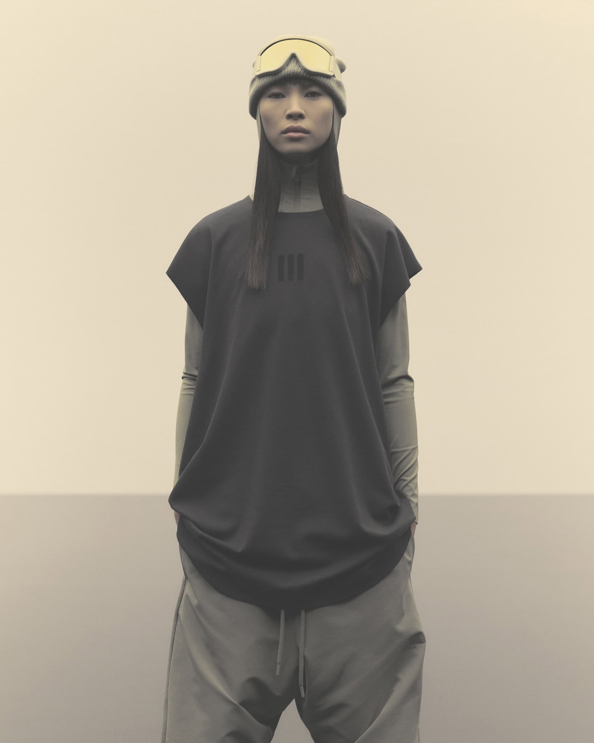 An Asian woman stands centered, wearing ski goggles on her head, a black shirt, and grey pants against a plain, neutral background.
