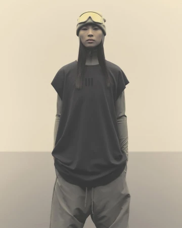 An Asian woman stands centered, wearing ski goggles on her head, a black shirt, and grey pants against a plain, neutral background.