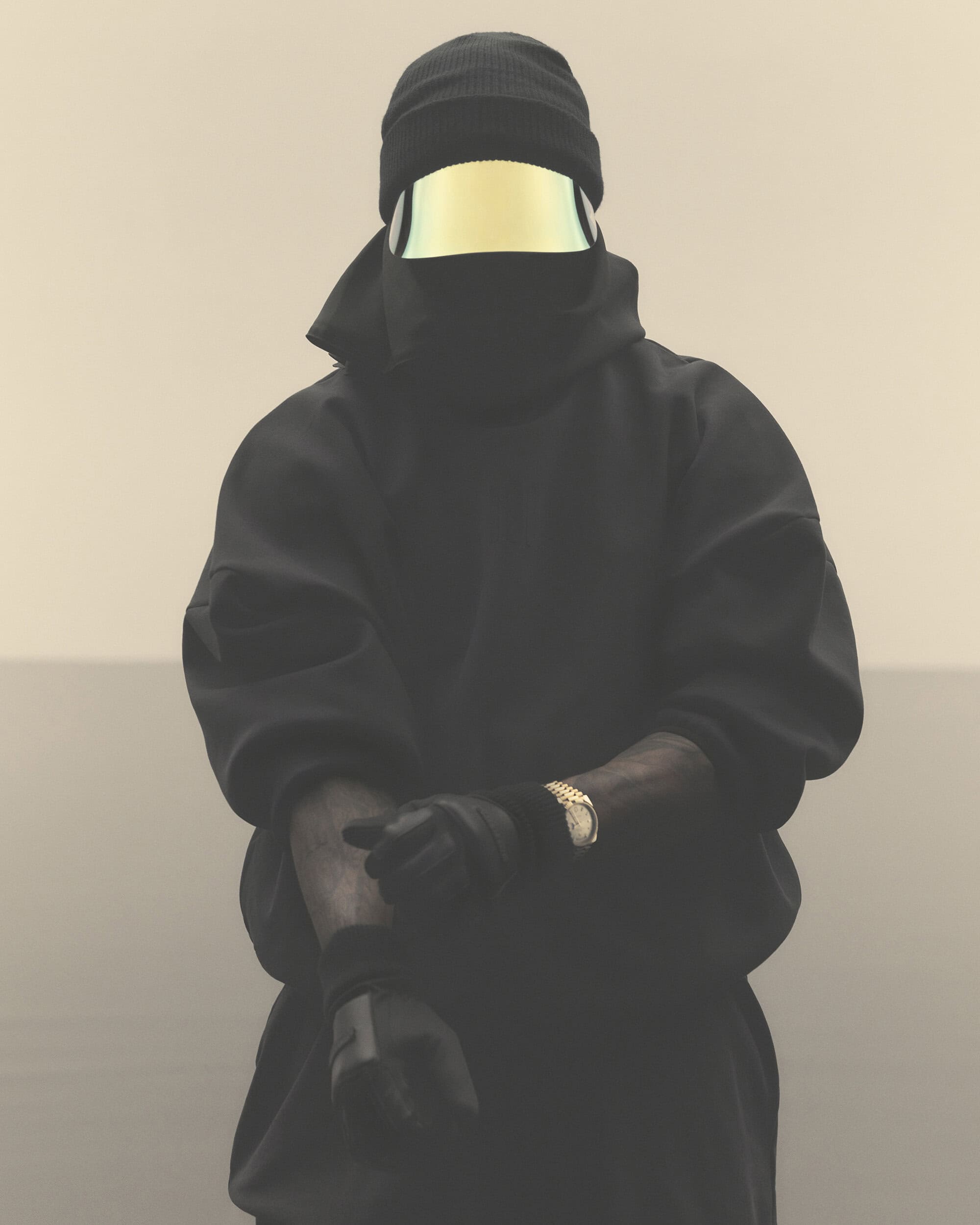 Person in black balaclava and gold visor, close-up studio shot on neutral backdrop, wearing a black hooded sweatshirt and gold watch.