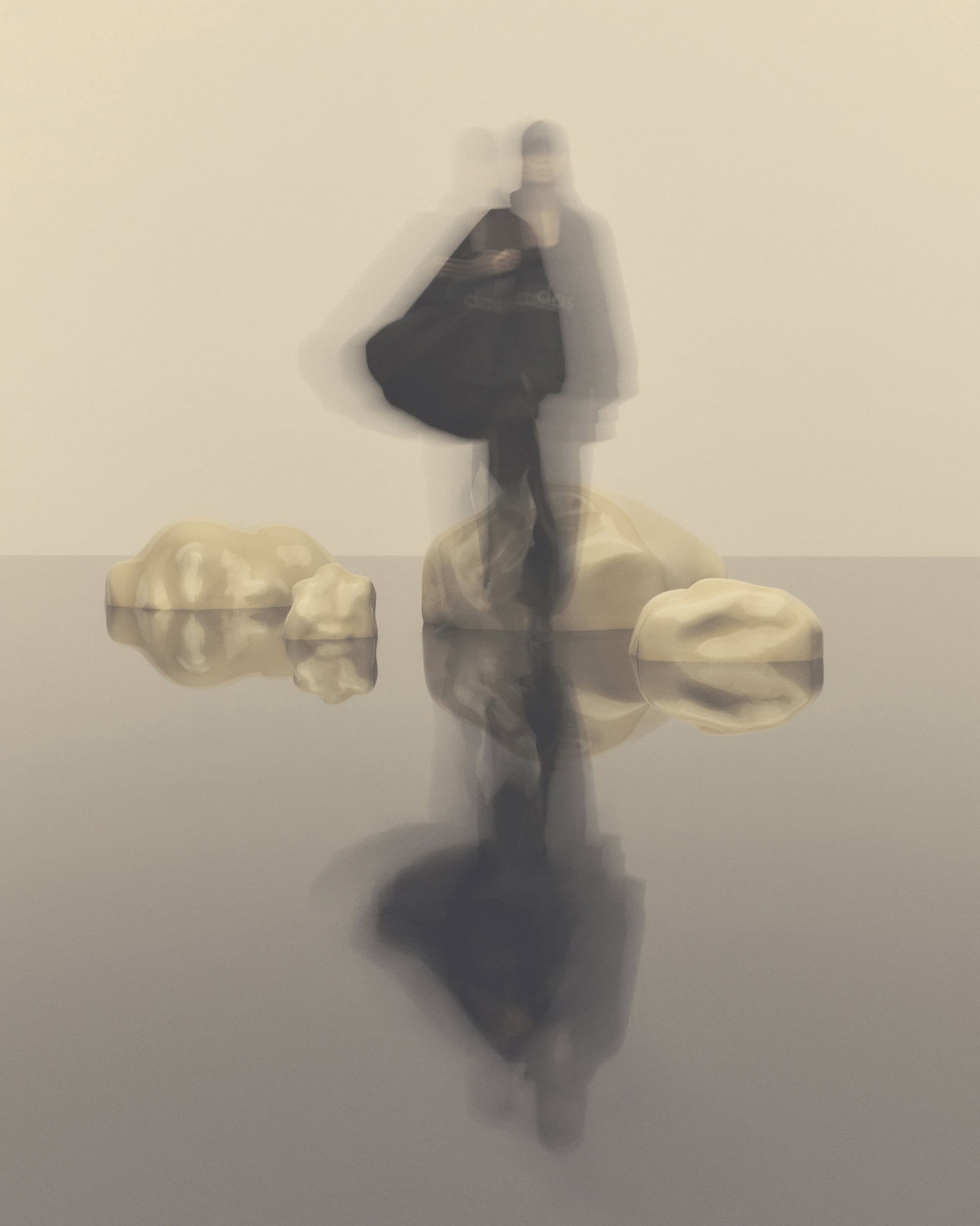 Abstract fashion shot: blurred figure in dark coat reflected on water, standing on rocks against a neutral backdrop.