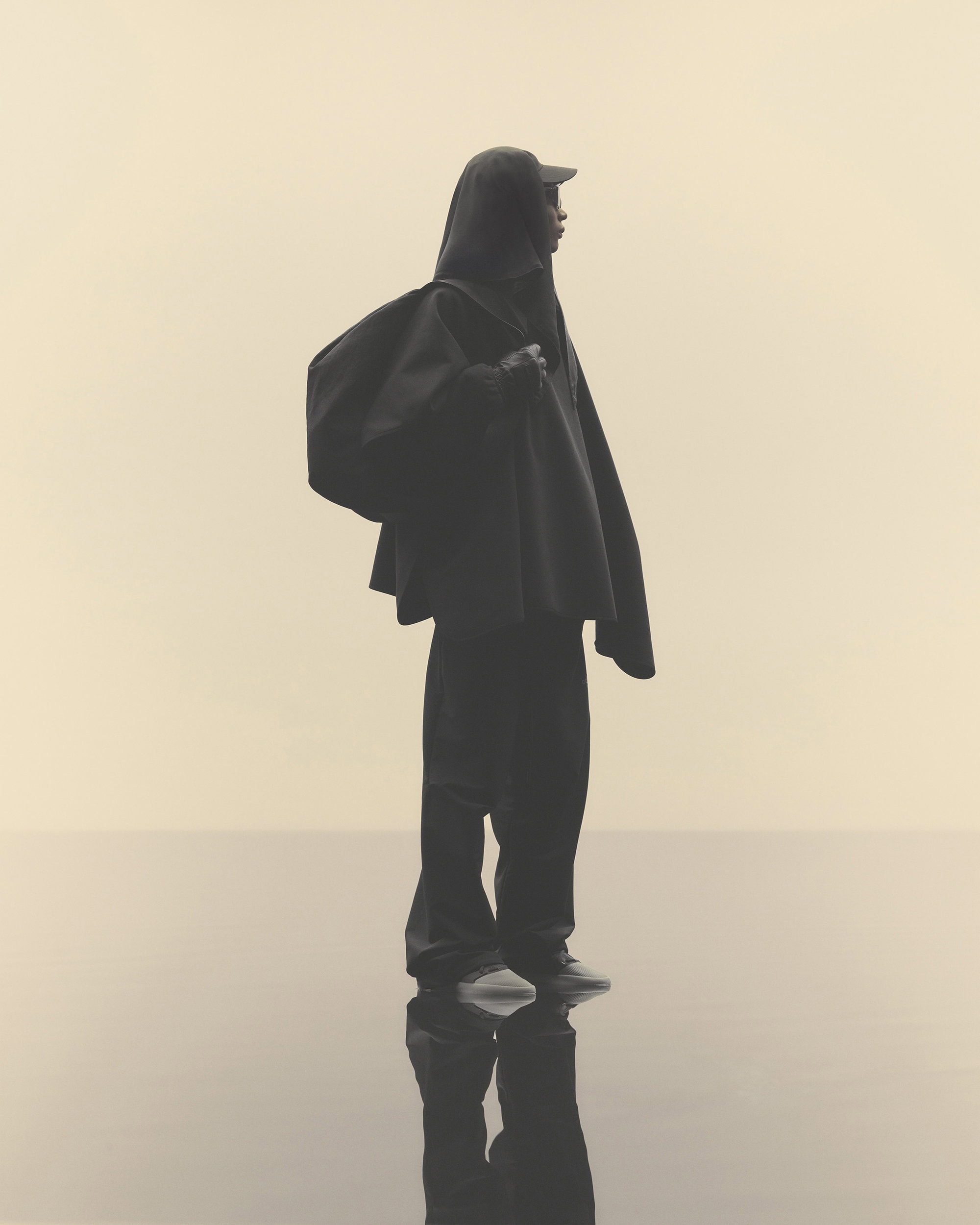 Person in dark hooded poncho stands on reflective surface against pale background in fashion studio lighting, full length view.