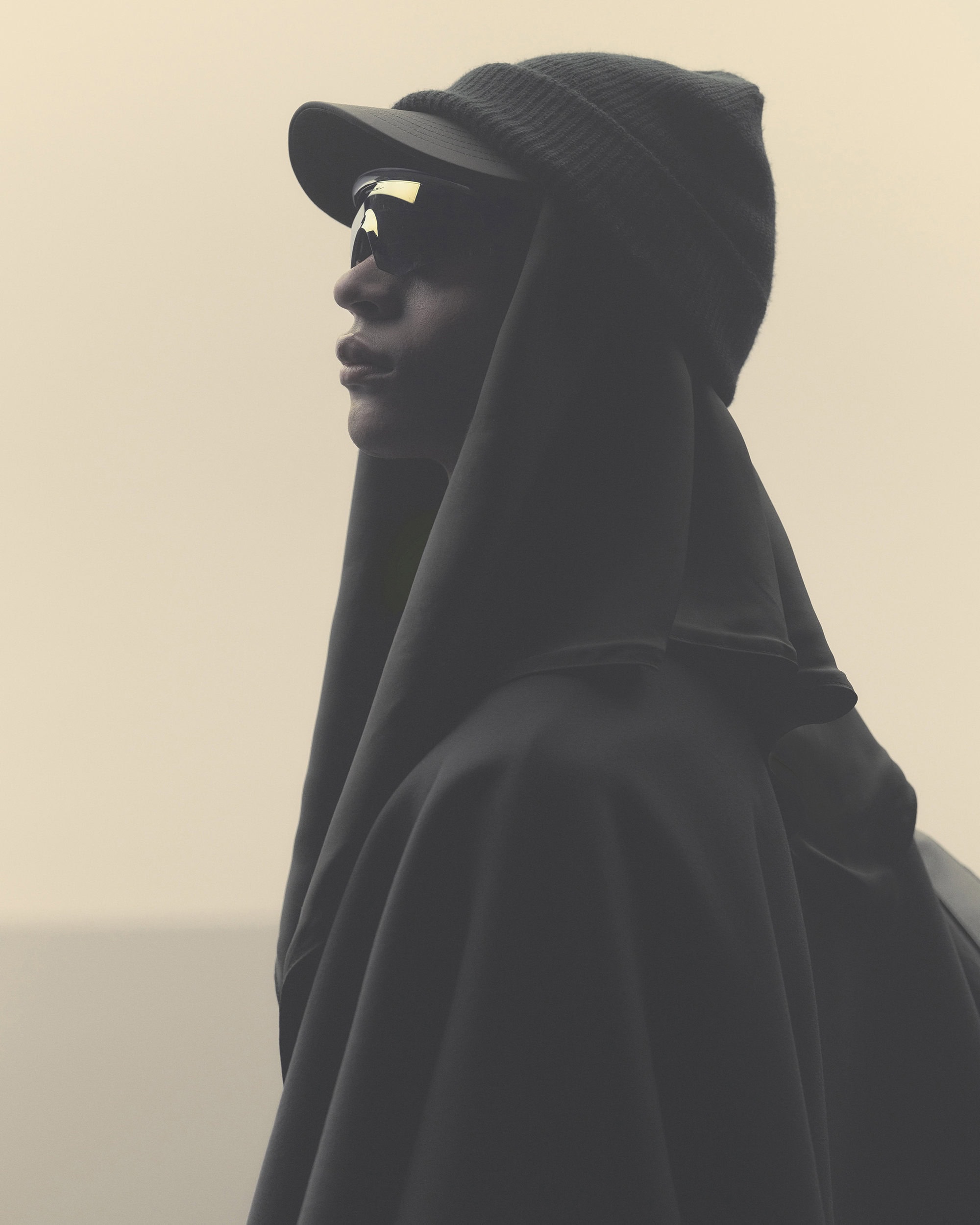 Side view of a person in a black hooded cape, sunglasses, and cap against a pale background, emphasizing dark, enveloping fashion.