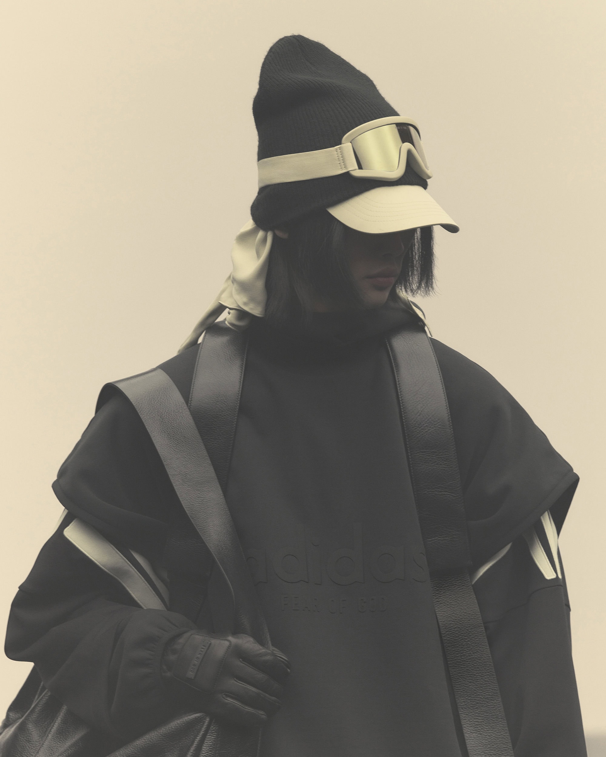 Person wearing a black knit hat with goggles and a cape-like coat, standing against a light background in a fashion-editorial style photograph.