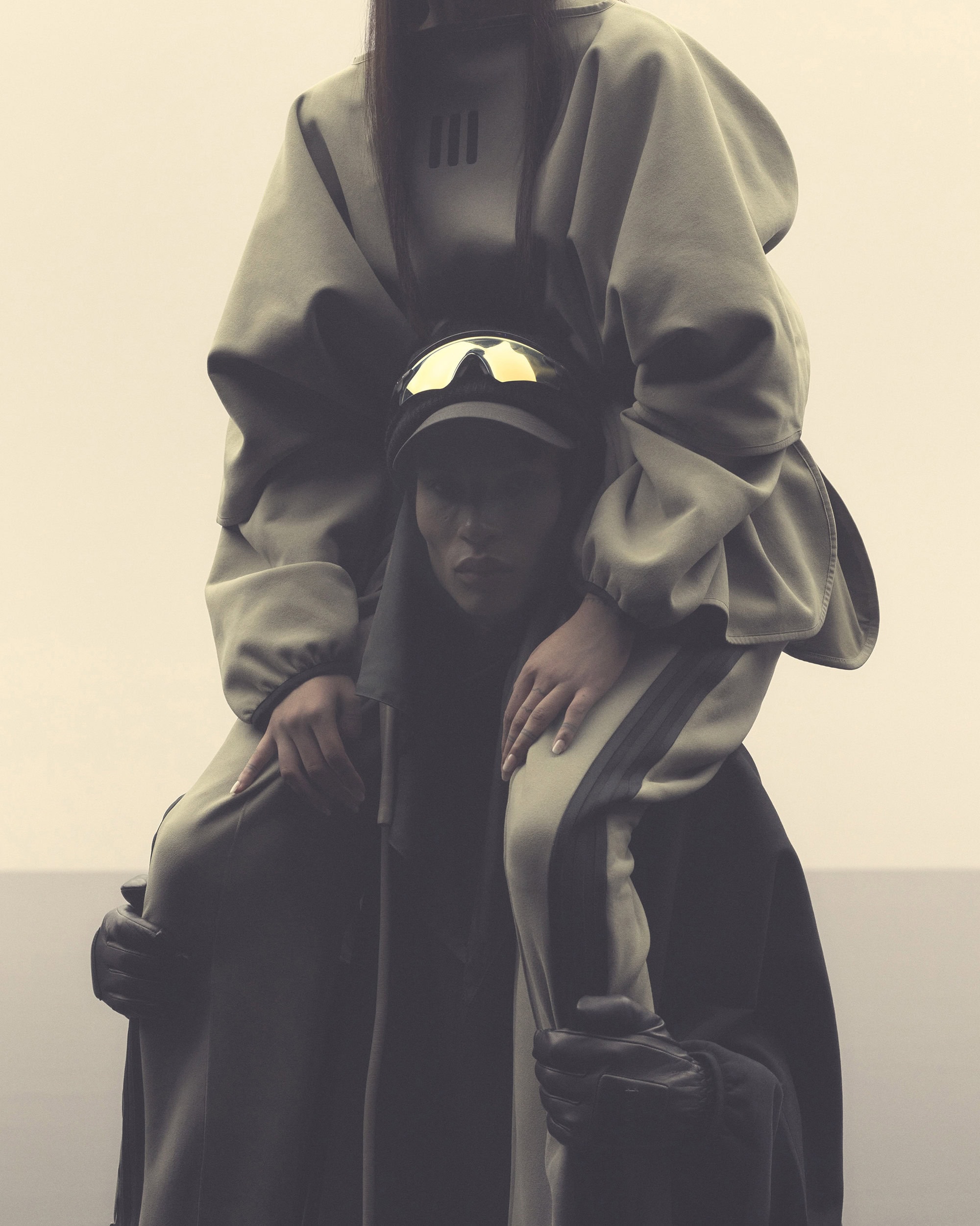 Two people wearing matching olive green athletic tracksuits are standing in front of a neutral-colored horizon with an overcast sky.