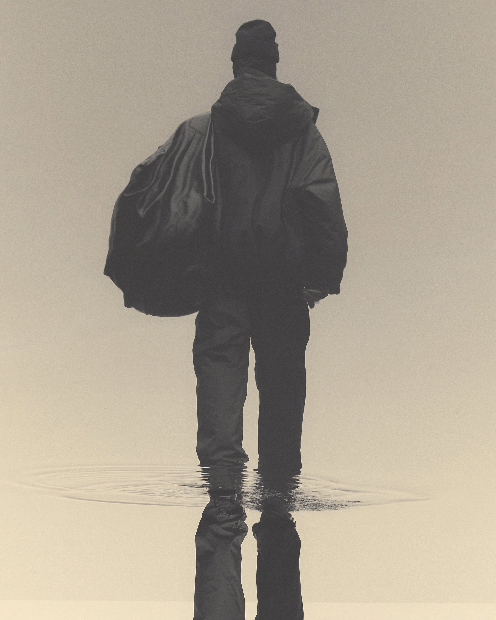 A person wearing a jacket and hat walks through shallow water, creating ripples. The scene is overcast and rendered in desaturated tones.