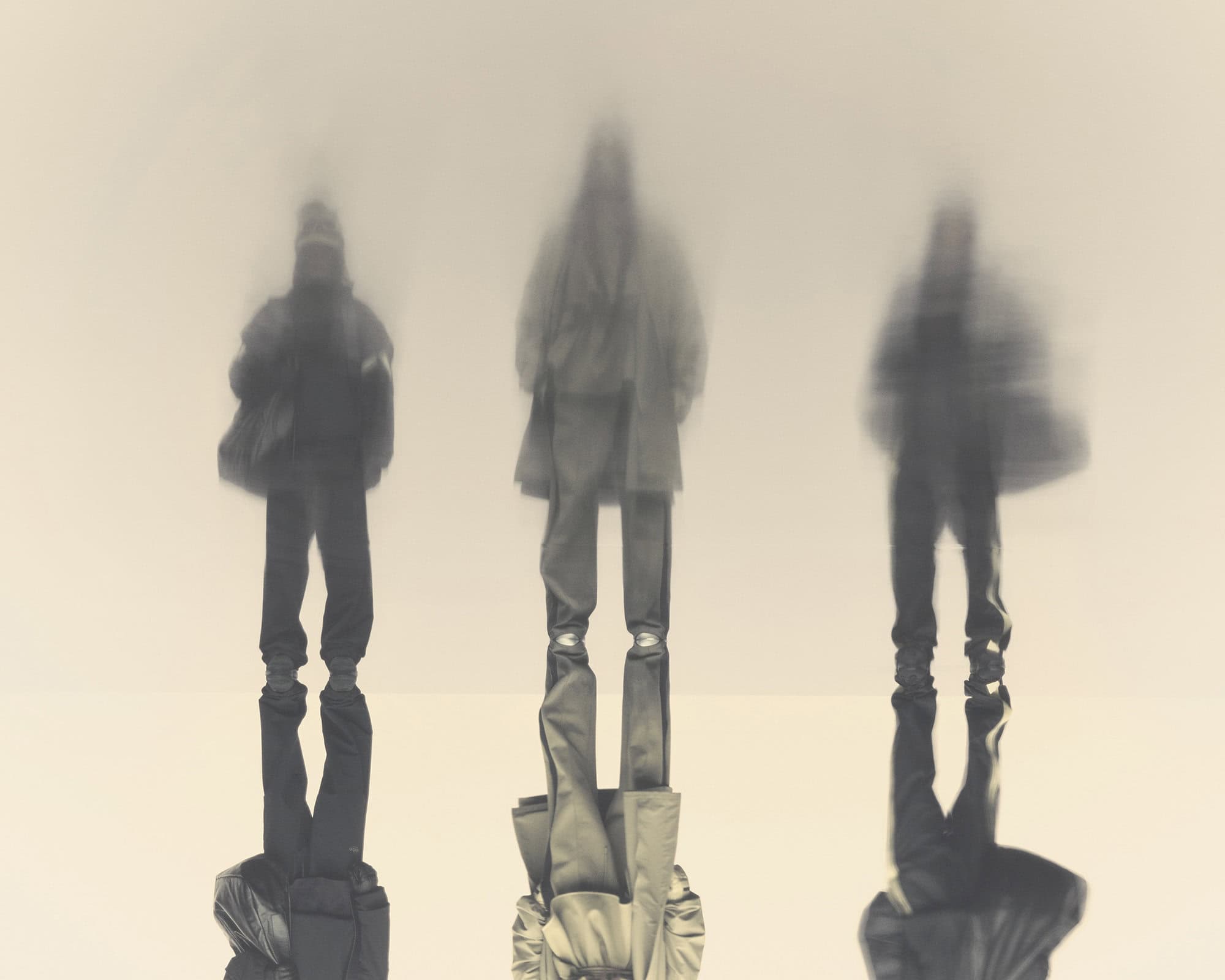 Three blurred figures wearing coats and trousers, standing outdoors, abstract appearance, desaturated colour palette.